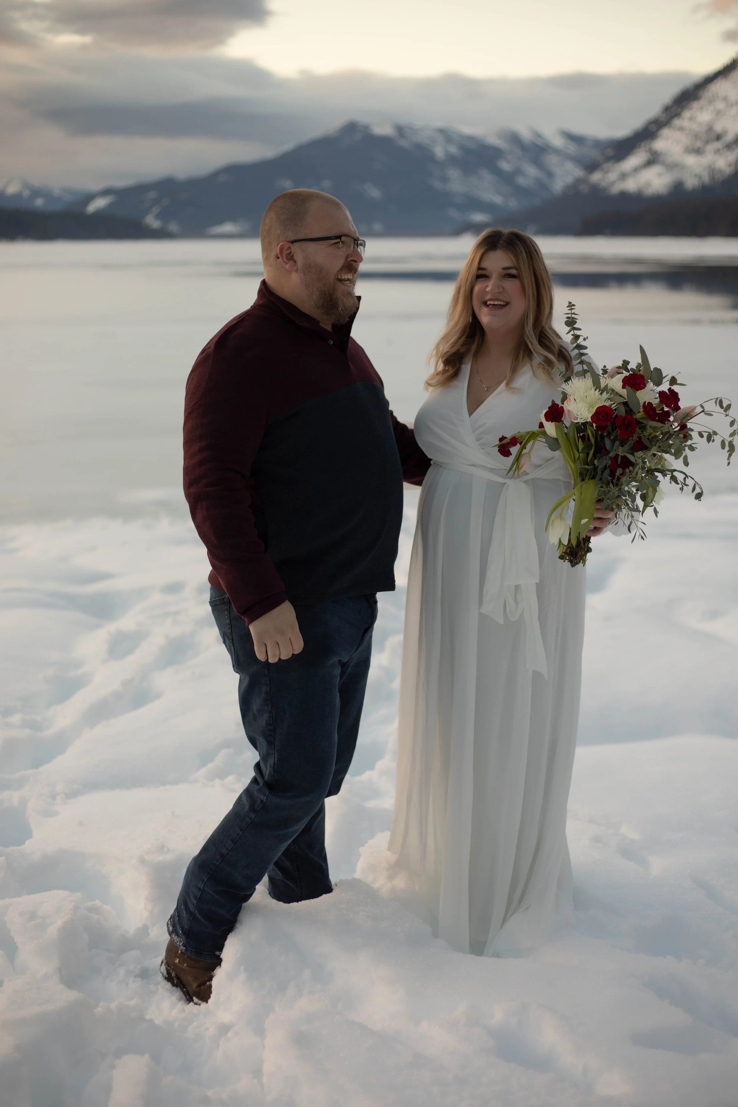 A couple stands together outdoors on snow, with mountains and a cloudy sky in the background. The woman wears a white dress and holds a bouquet, while the man wears a dark sweater and jeans, smiling at each other.