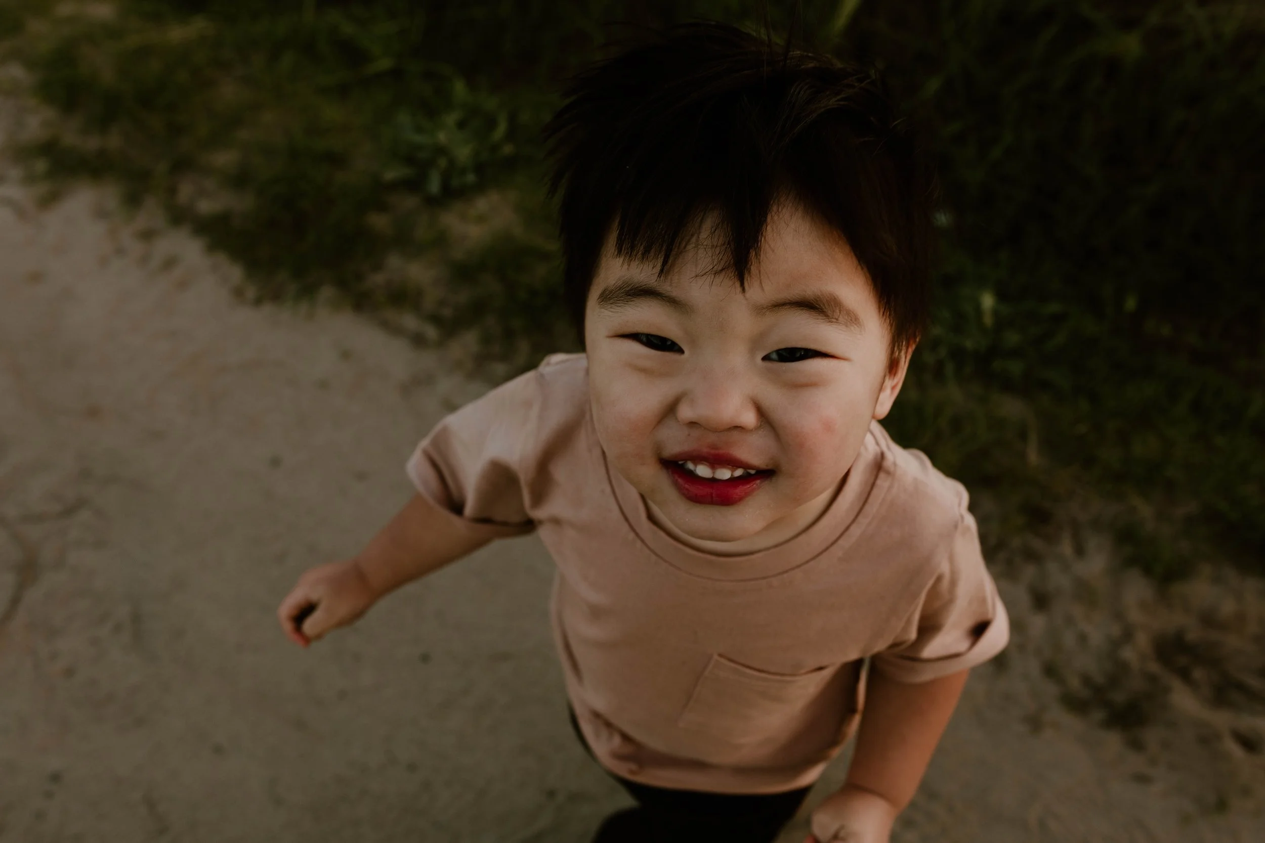 Young boy with short dark hair, smiling up at the camera, wearing a beige t-shirt, standing outdoors on a dirt path with grass and plants in the background.