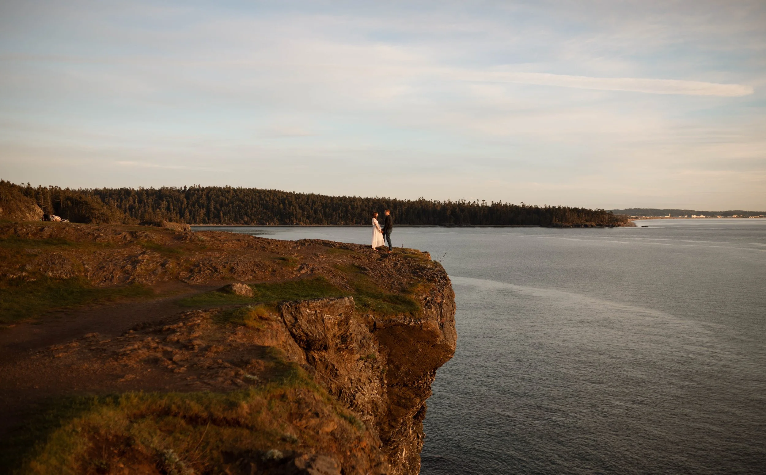 A couple is standing on the edge of a cliff overlooking a body of water, with a forest in the background and a partly cloudy sky.
