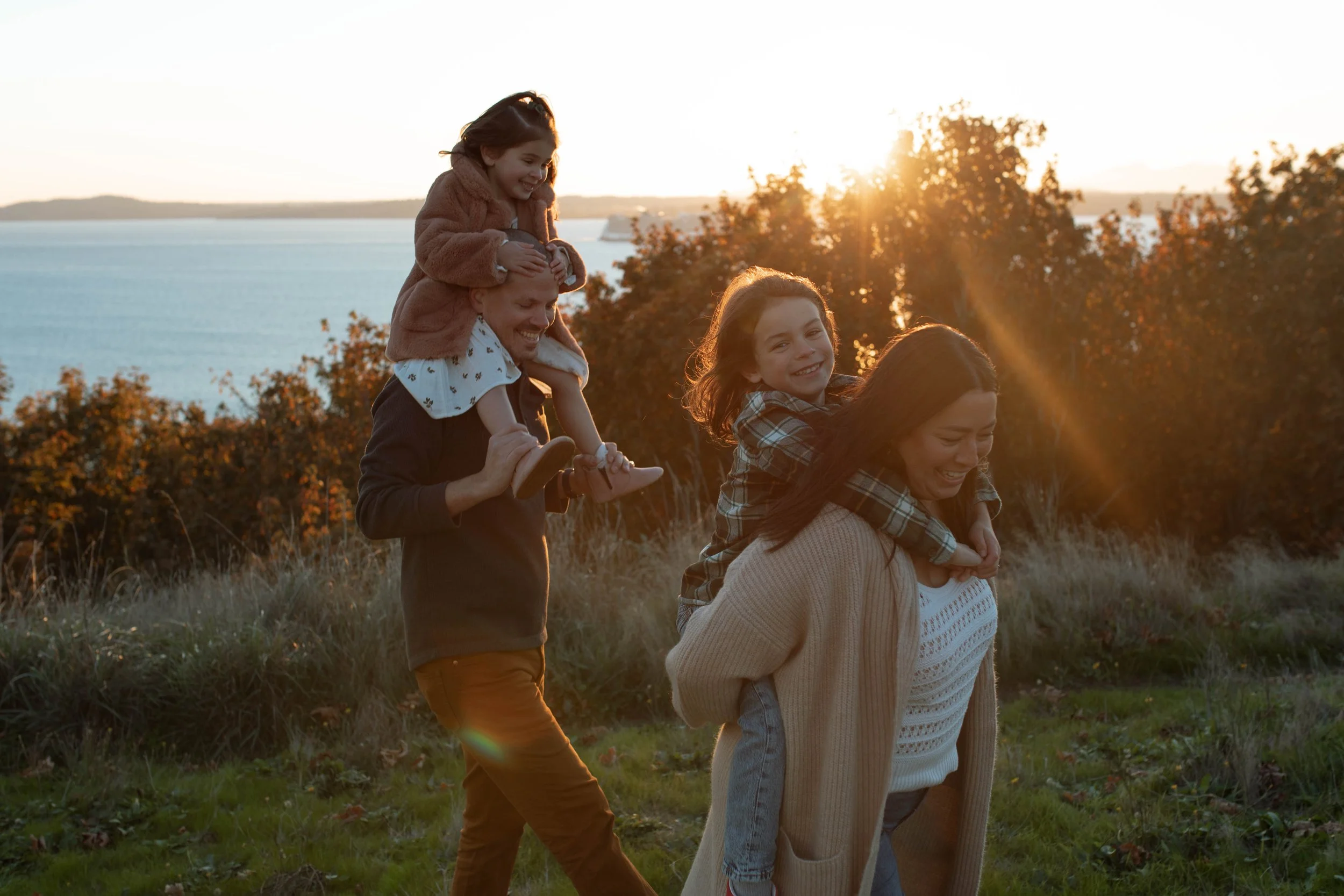 A family of four enjoying a walk outdoors at sunset, with two children riding on their parents' shoulders.