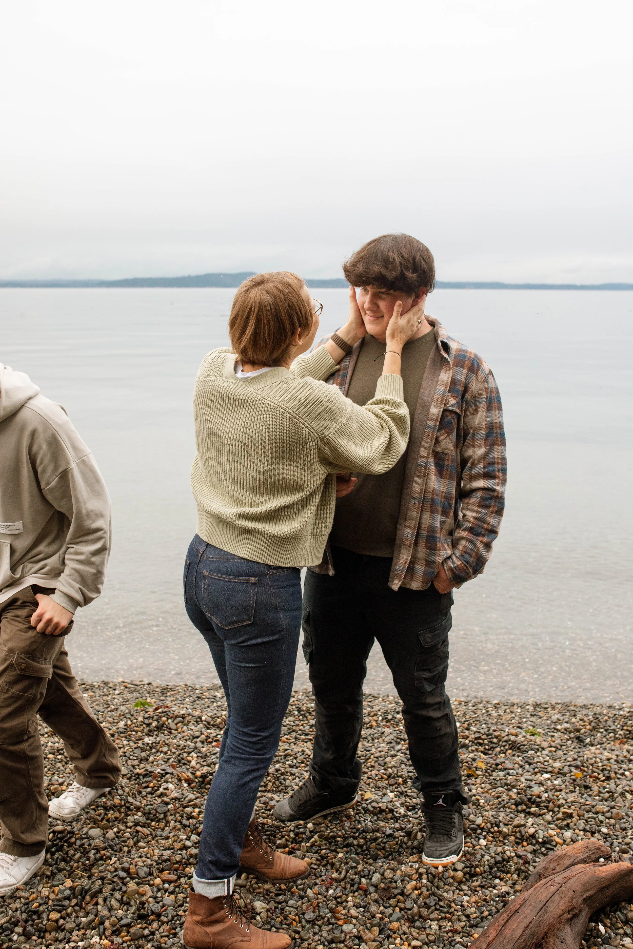 A woman and a young man stand on a pebbled beach by a body of water, smiling and touching each other's faces affectionately.