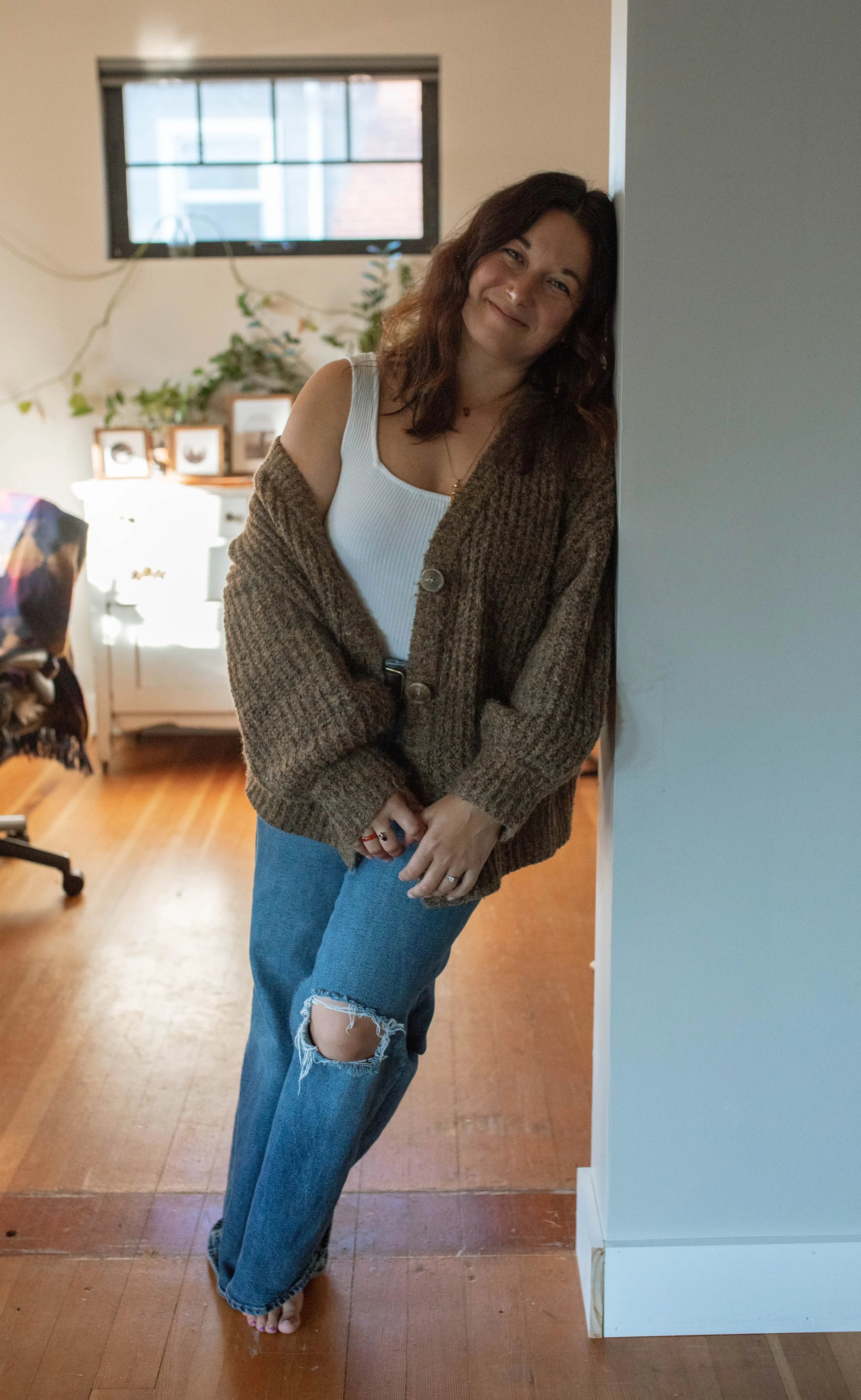 A woman leaning against a wall in a cozy, well-lit room, smiling at the camera, wearing a white tank top, a brown cardigan, and ripped jeans.