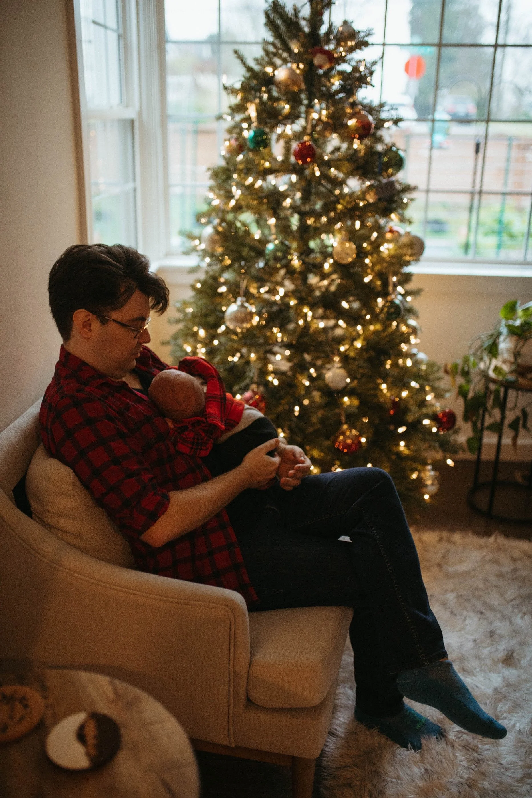 A man sitting on a beige armchair holding a baby near a decorated Christmas tree in a living room.