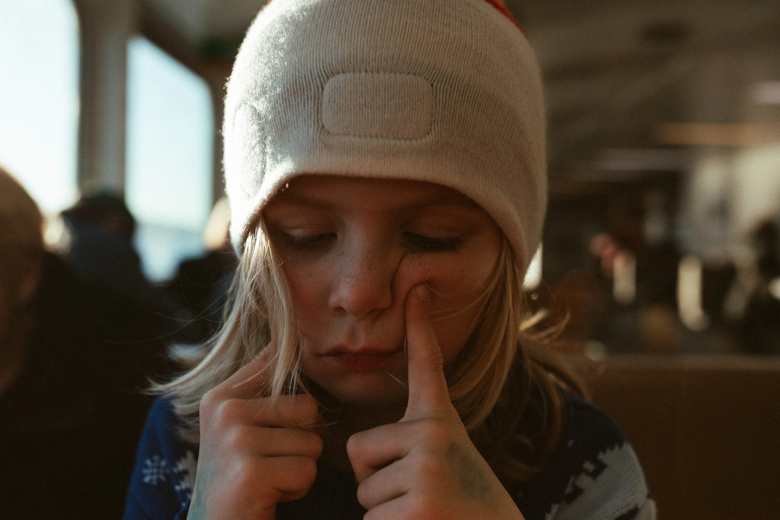 A young woman with blonde hair in a white knit beanie with a square patch, sitting indoors with blurred figures in the background. She is touching her face with one finger, with her eyes closed and a thoughtful expression.