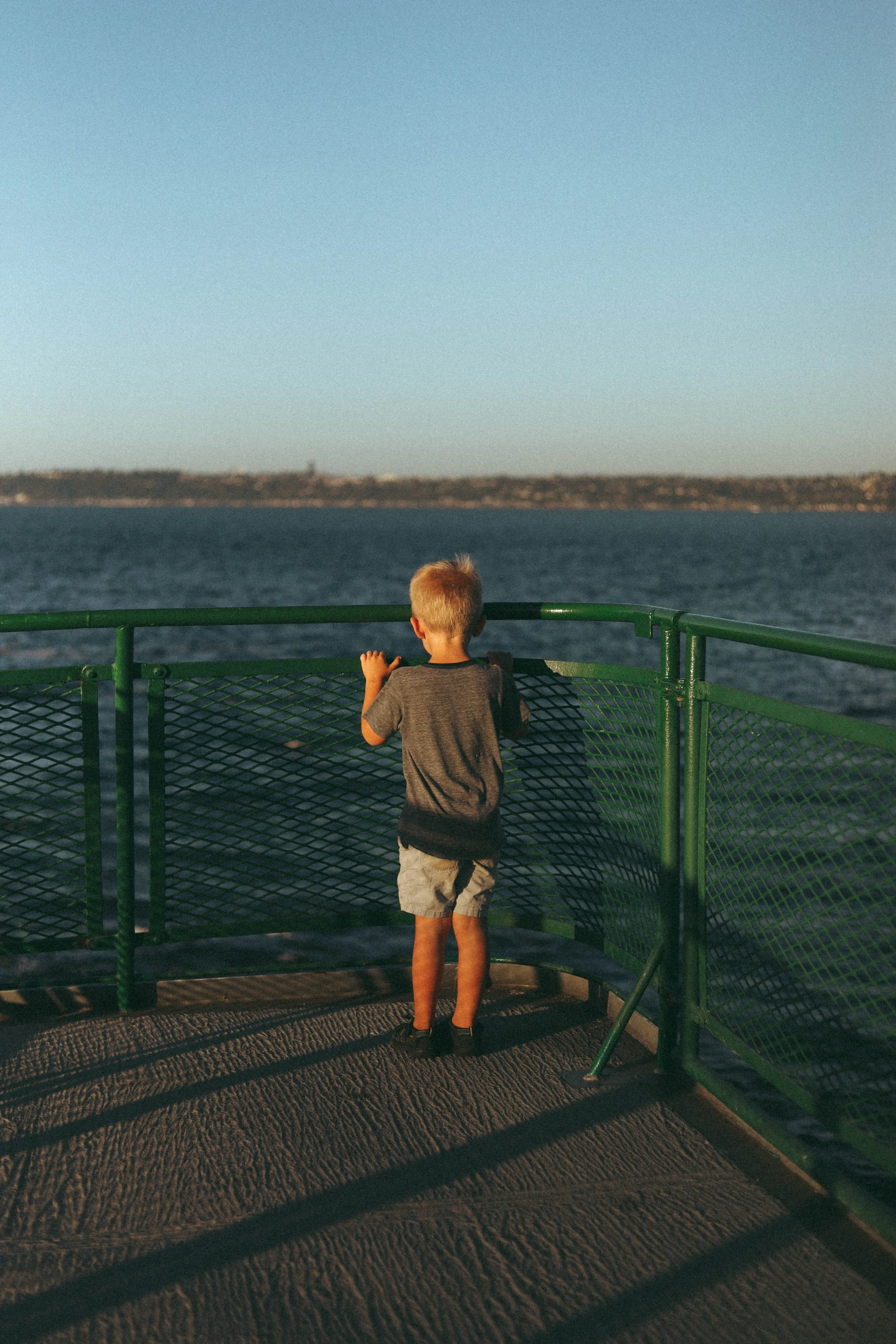 A young boy with blonde hair wearing a grey T-shirt and shorts looking out over a body of water from a green metal railing on a boat.