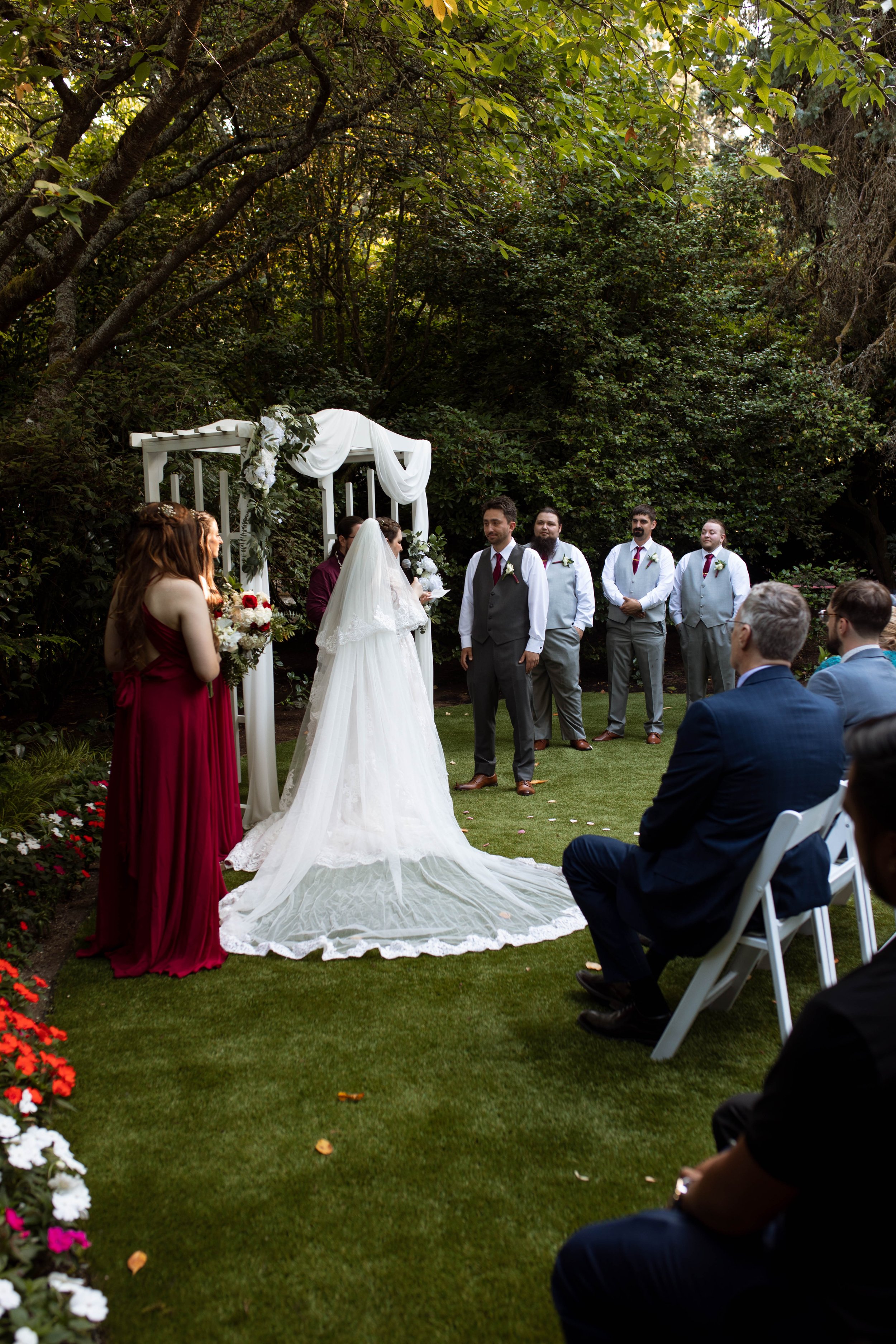 A wedding ceremony taking place outdoors in a garden with greenery. The bride in a white wedding gown and veil stands under a white arch decorated with flowers and draped fabric. The groom and groomsmen, dressed in suits, stand nearby. The bridesmaid
