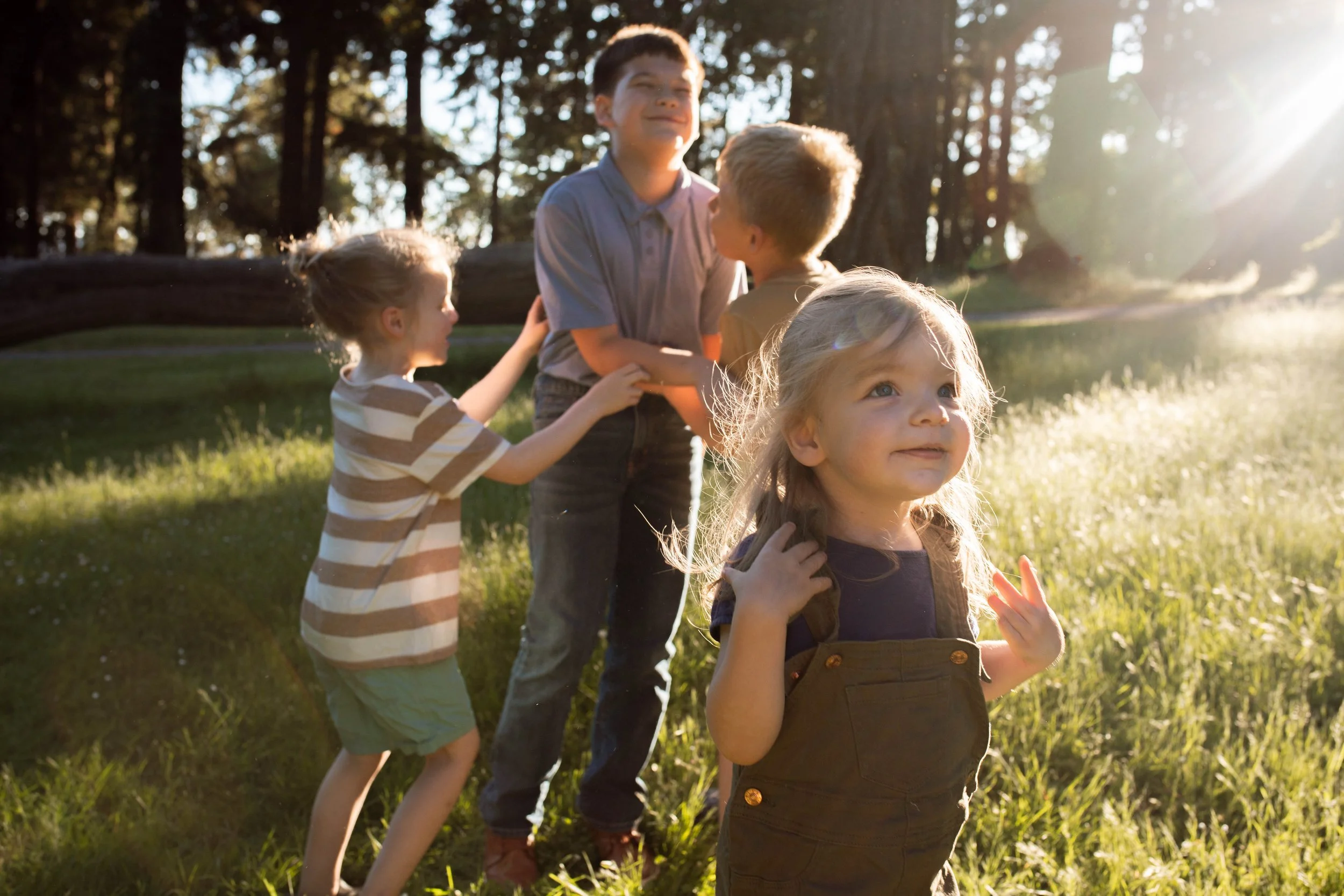Four children play together in a sunlit forest clearing, with a large fallen tree in the background.
