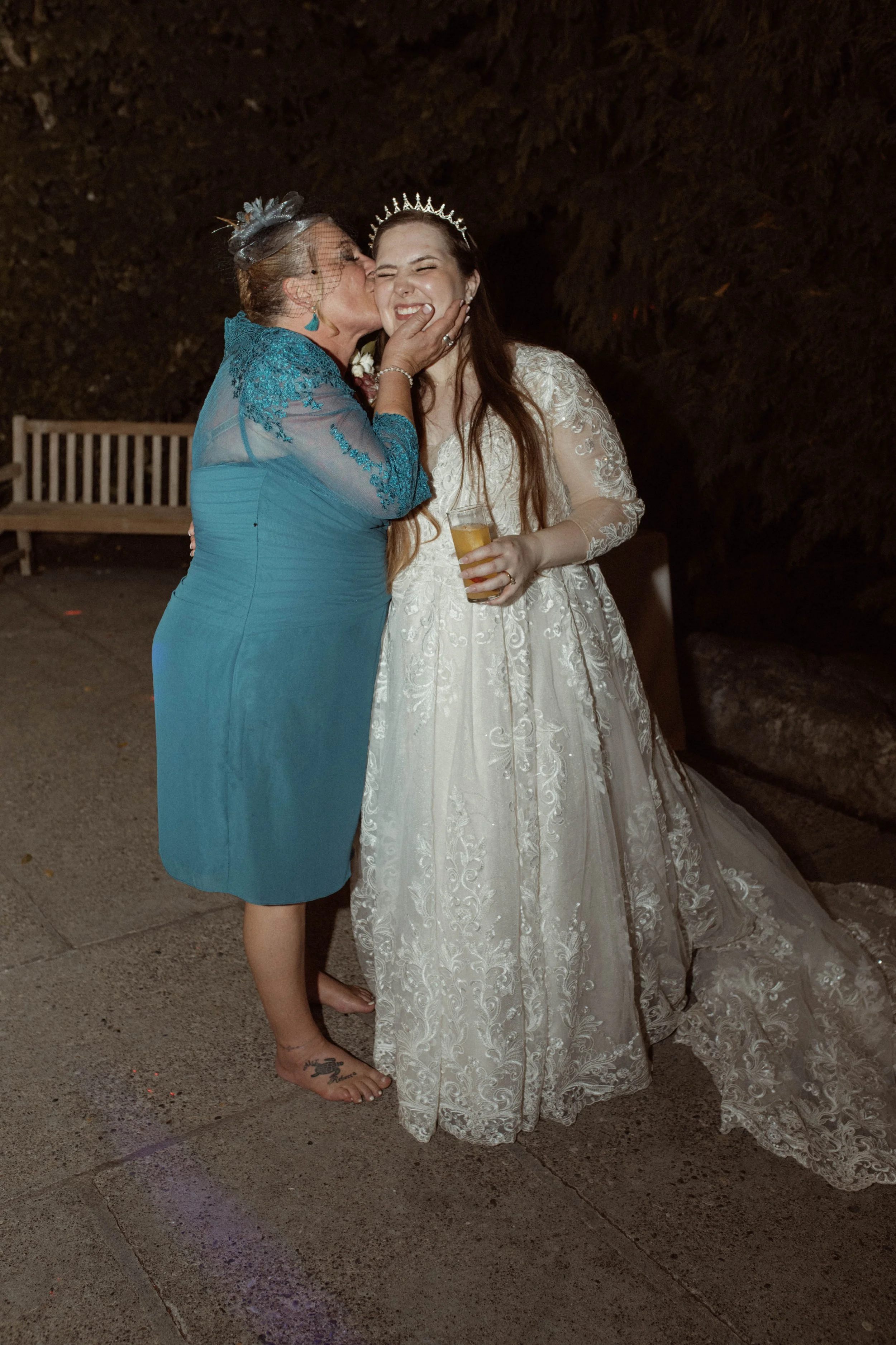 A woman in a blue dress is kissing a young woman in a white wedding gown on the cheek, both are smiling. The young woman is holding a drink and wearing a tiara, while the older woman is wearing a fascinator hat.