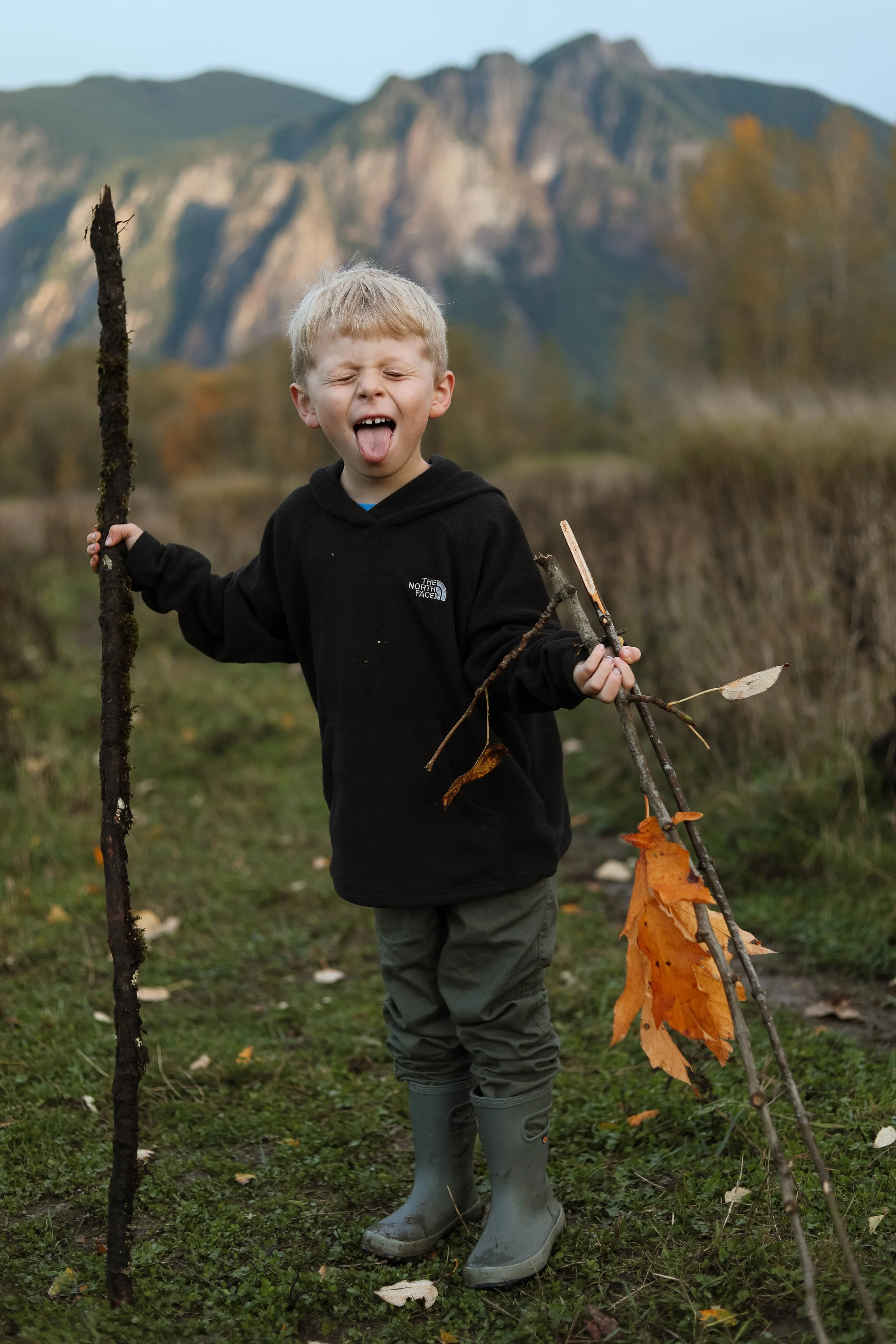 A young boy outdoors in a grassy area holding a stick and a small branch with autumn leaves, making a funny face with his tongue out and eyes closed. He is dressed in a black hoodie and gray rain boots, with mountains in the background.