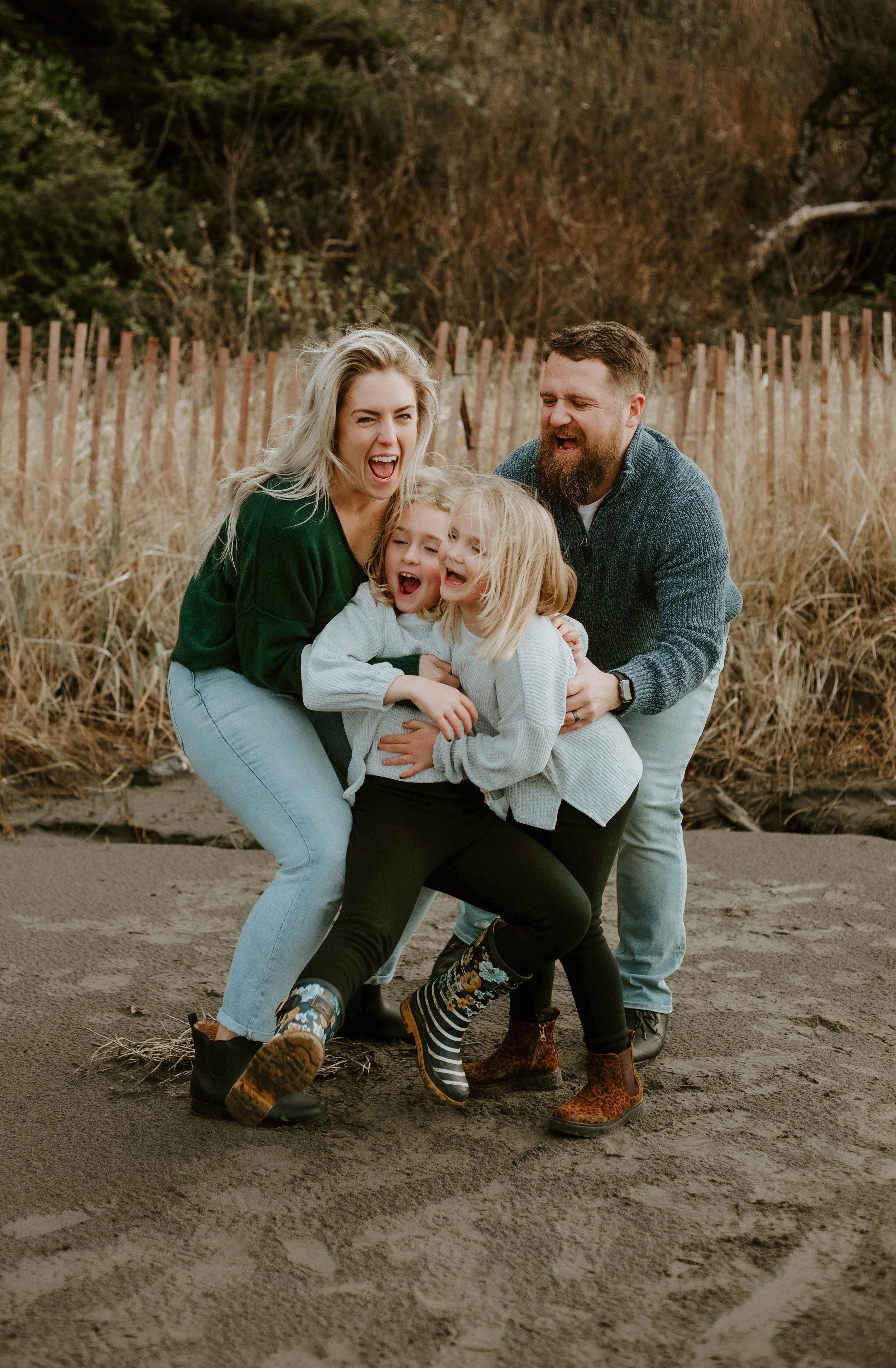 A family of four, two adults and two children, laughing and hugging on a sandy beach with tall grass and a wooden fence in the background.