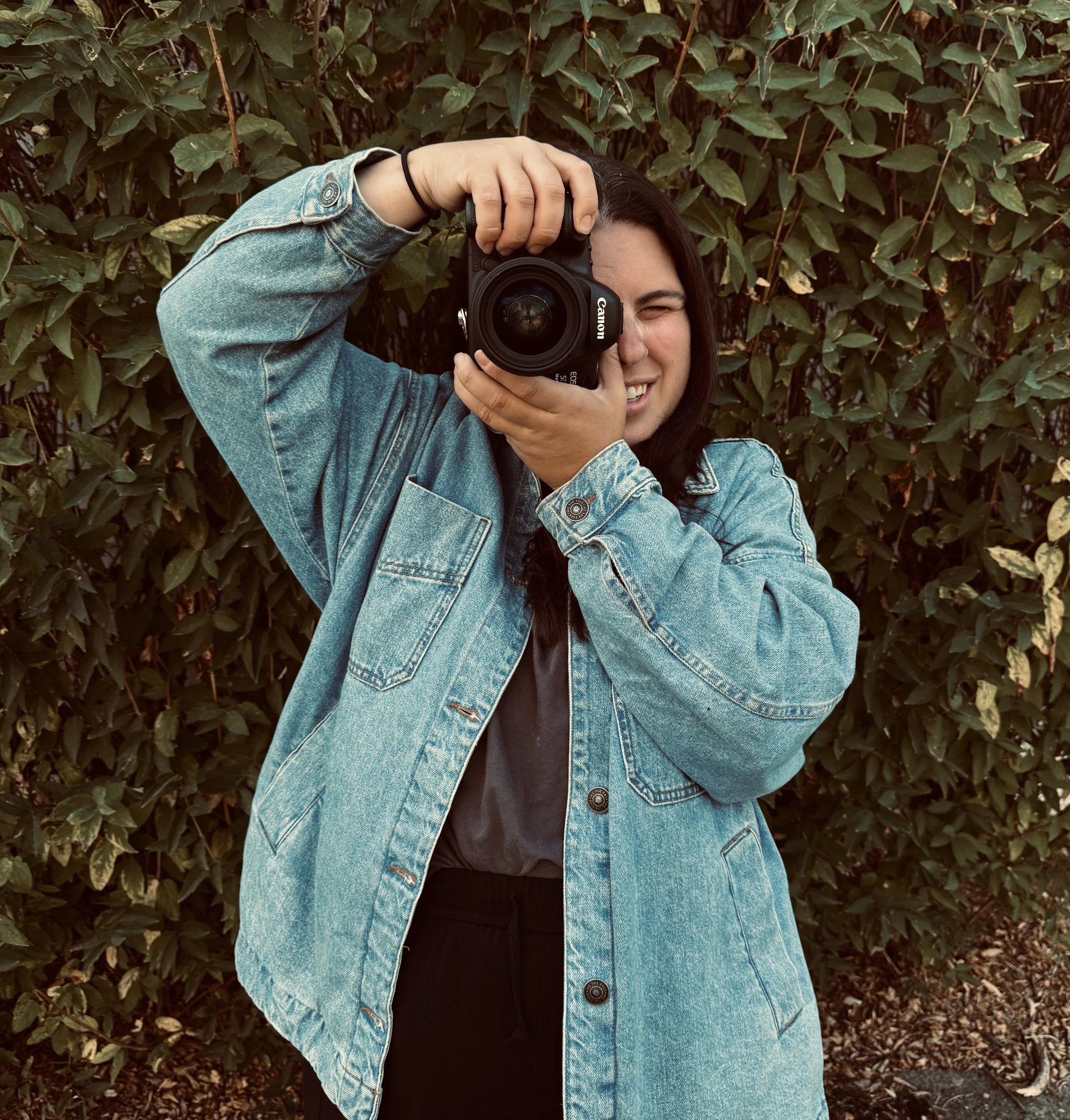 A woman with dark hair wearing a denim jacket is taking a photo with a camera, standing in front of green leafy bushes.