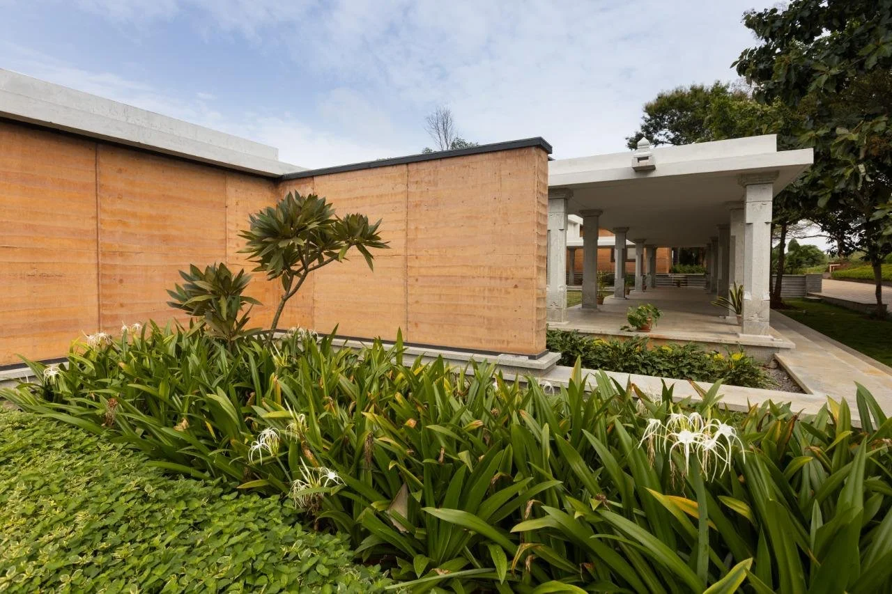 A modern building with a covered walkway supported by stone columns, surrounded by green plants and trees, with a partly cloudy sky overhead.