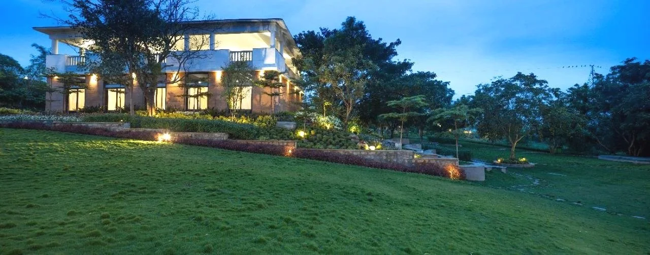 A modern house with large windows and a balcony, illuminated at dusk, surrounded by well-maintained lawn, trees, and landscaped garden with pathway lighting.