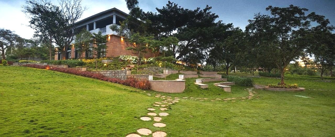 A hilly landscaped garden with a winding stone path leading to a modern house with a spacious balcony, surrounded by trees and plants, during dusk with lights on.