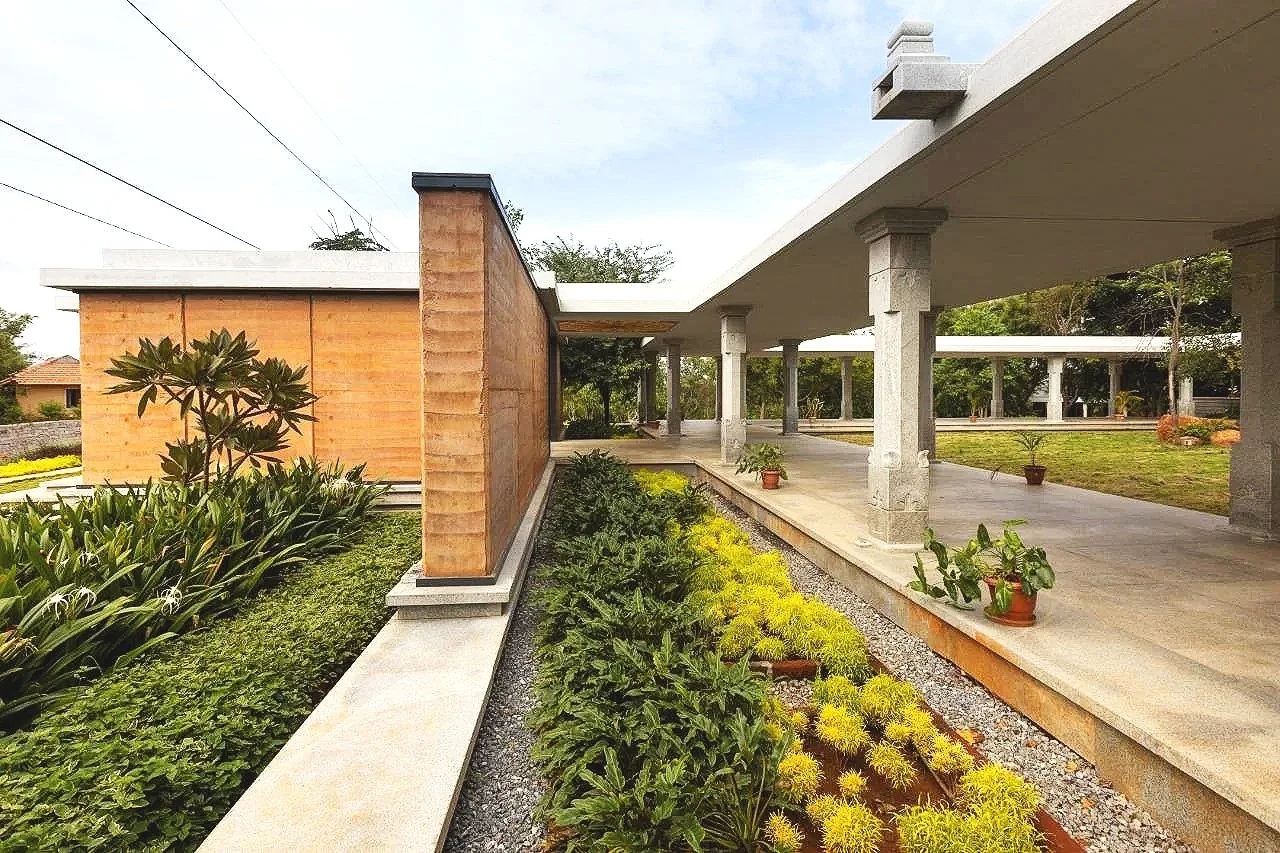 Modern house with open walkway, stone columns, and landscaped garden with potted plants and various shrubs.
