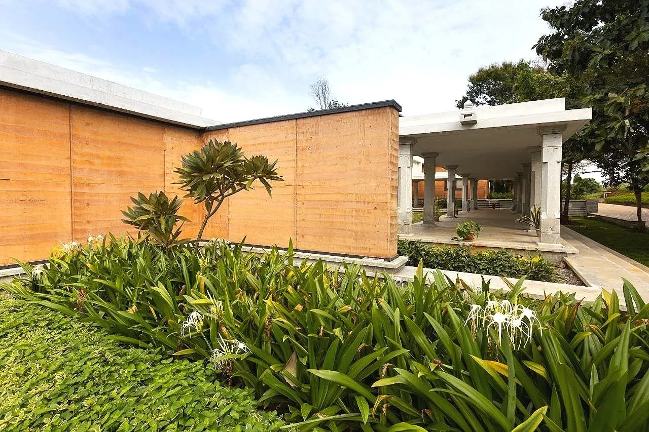 A modern building with a covered walkway supported by stone columns, surrounded by green plants and trees, with a partly cloudy sky overhead.