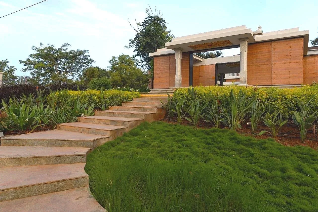 A modern house with a flat roof and concrete and wood construction, surrounded by landscaped greenery, with a stone walkway leading up to it.