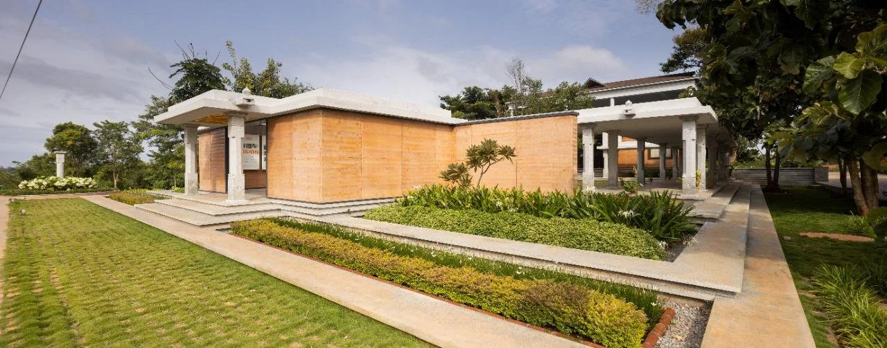 Modern building with brick walls, white concrete pillars, and landscaped garden with green grass, shrubs, and trees.