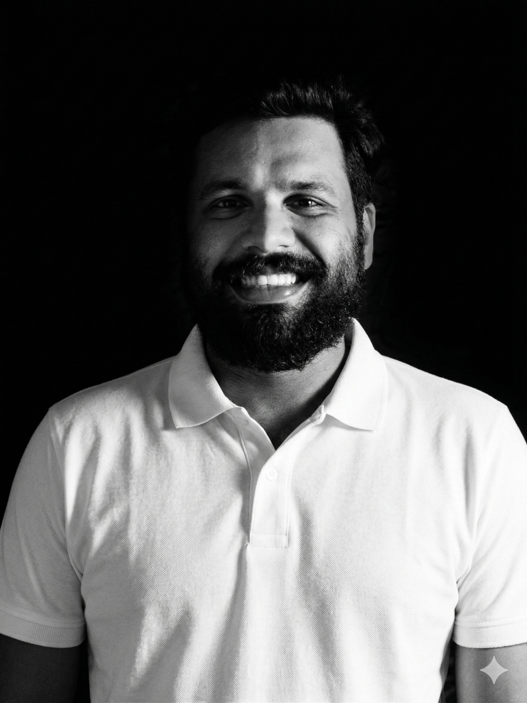 A black and white portrait of a smiling man with a beard, wearing a collared shirt, against a dark background.