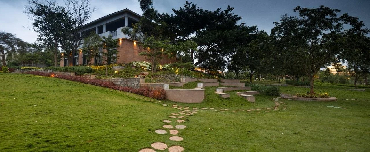 A hilly landscaped garden with a winding stone path leading to a modern house with a spacious balcony, surrounded by trees and plants, during dusk with lights on.
