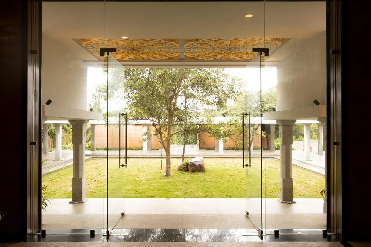 Glass doors open to a courtyard with a tree, grass, rocks, and surrounding columns.