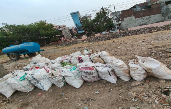 A row of white sacks filled with debris or materials, lined up on a dirt ground in an urban area with buildings and trees in the background.
