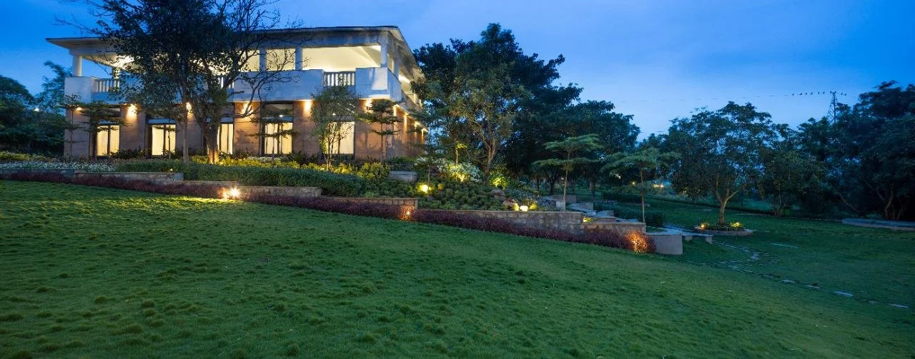 A modern house with large windows and a balcony, illuminated at dusk, surrounded by well-maintained lawn, trees, and landscaped garden with pathway lighting.