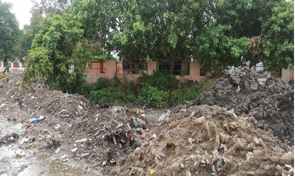 A construction site with piles of dirt and rocks, scattered trash, and a partially flooded area with a backdrop of green trees and a pink wall with iron railings.