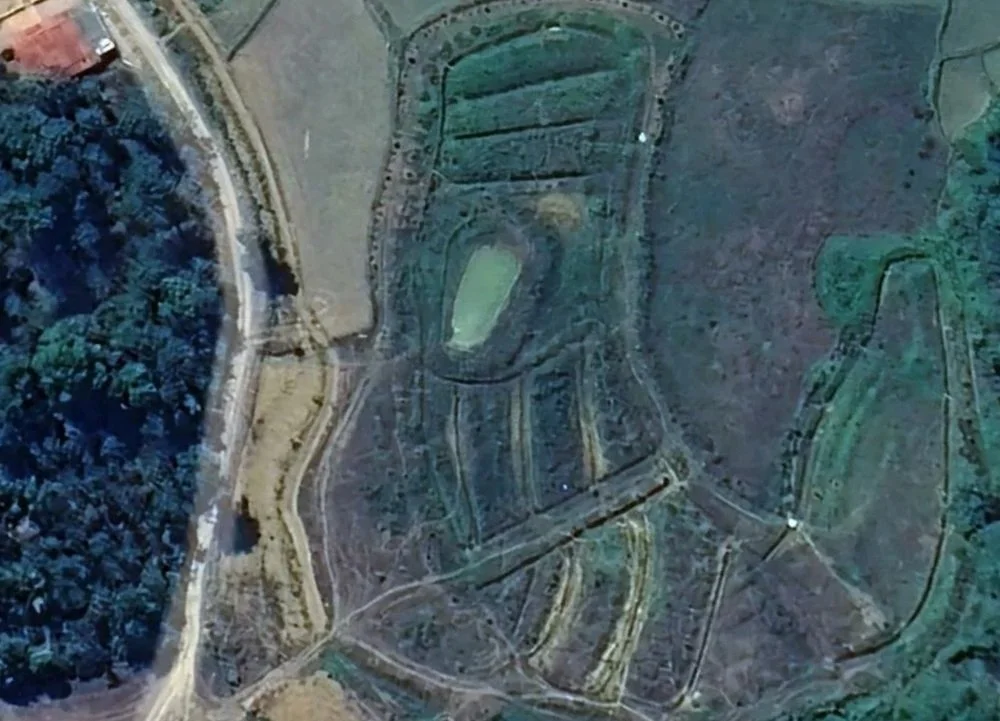 An aerial view of a landscape with a small oval-shaped pond, surrounding roads, and patches of green and brown land, possibly farmland or parks.