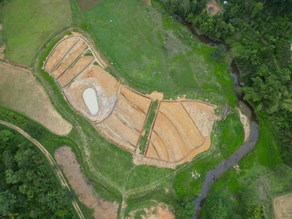 Aerial view of a construction site with terraced areas around a small water body, surrounded by green fields, trees, and a winding stream.