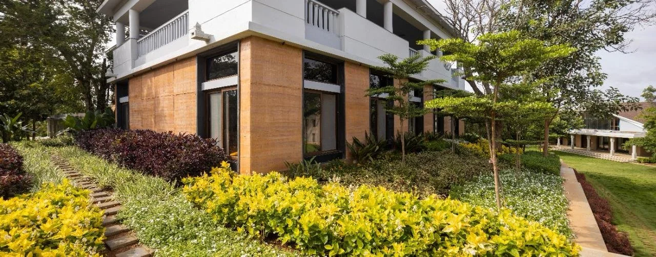 A modern building with large windows, surrounded by a landscaped garden with various green and yellow plants and trees.