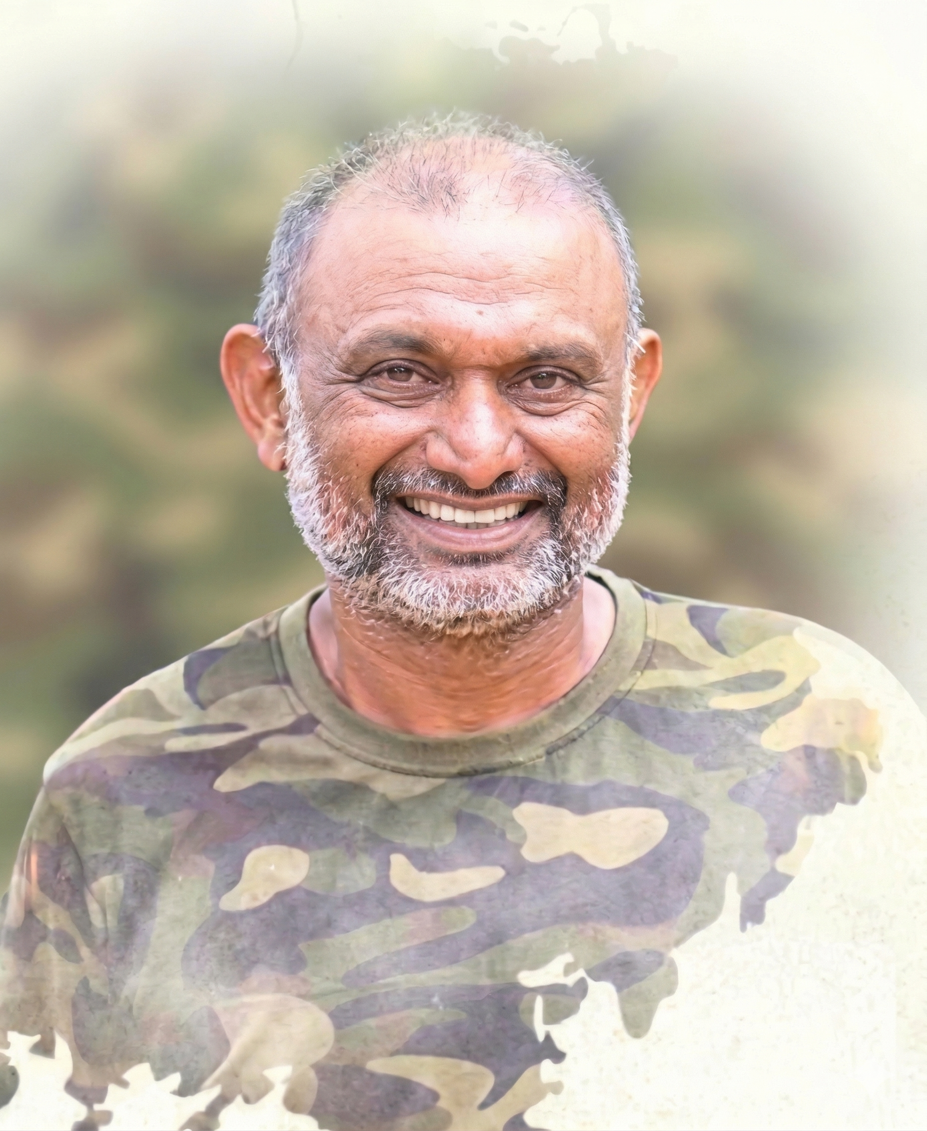 A smiling older man with a beard and short gray hair, wearing a camouflage shirt, outdoors with a blurred natural background.