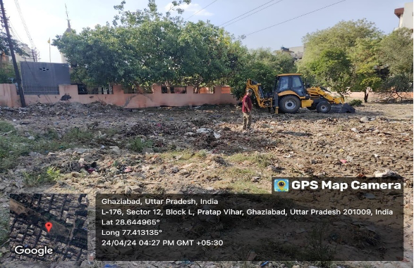 Empty lot with a yellow and black backhoe loader, a person standing nearby, and trees in the background. Overlay shows GPS coordinates and location details in Ghaziabad, Uttar Pradesh, India.