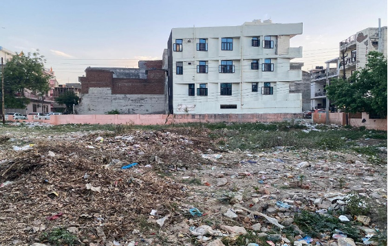 Empty lot with scattered trash and debris in urban area, multi-story buildings in the background.