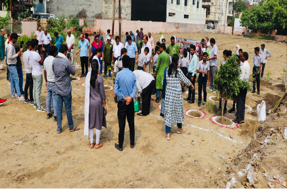 A group of people gathered outdoors on a dirt area, participating in a planting or environmental event, with some planting trees and others observing.