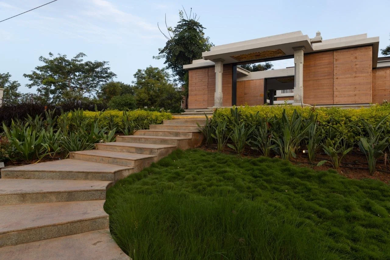 A modern house with a flat roof and concrete and wood construction, surrounded by landscaped greenery, with a stone walkway leading up to it.