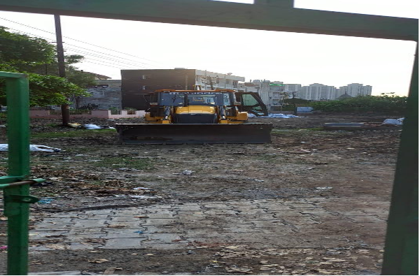 Construction site with a yellow bulldozer on dirt, seen through a green fence, with buildings in the background.