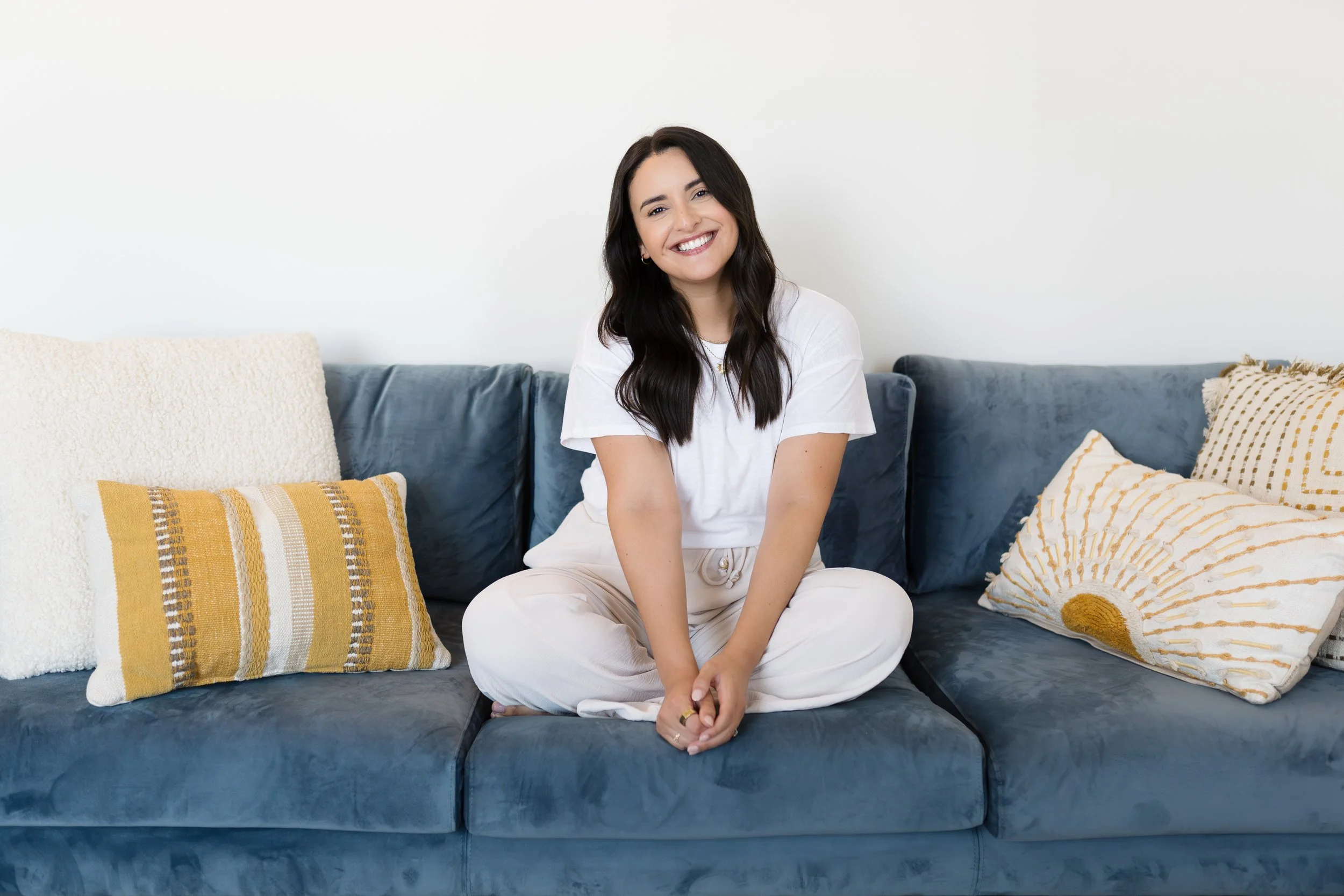A woman with long dark hair is sitting cross-legged on a blue velvet couch, smiling at the camera. She is wearing a white t-shirt and beige pants. There are decorative pillows on either side of her, one yellow and striped, and the other off-white with a sunburst pattern.