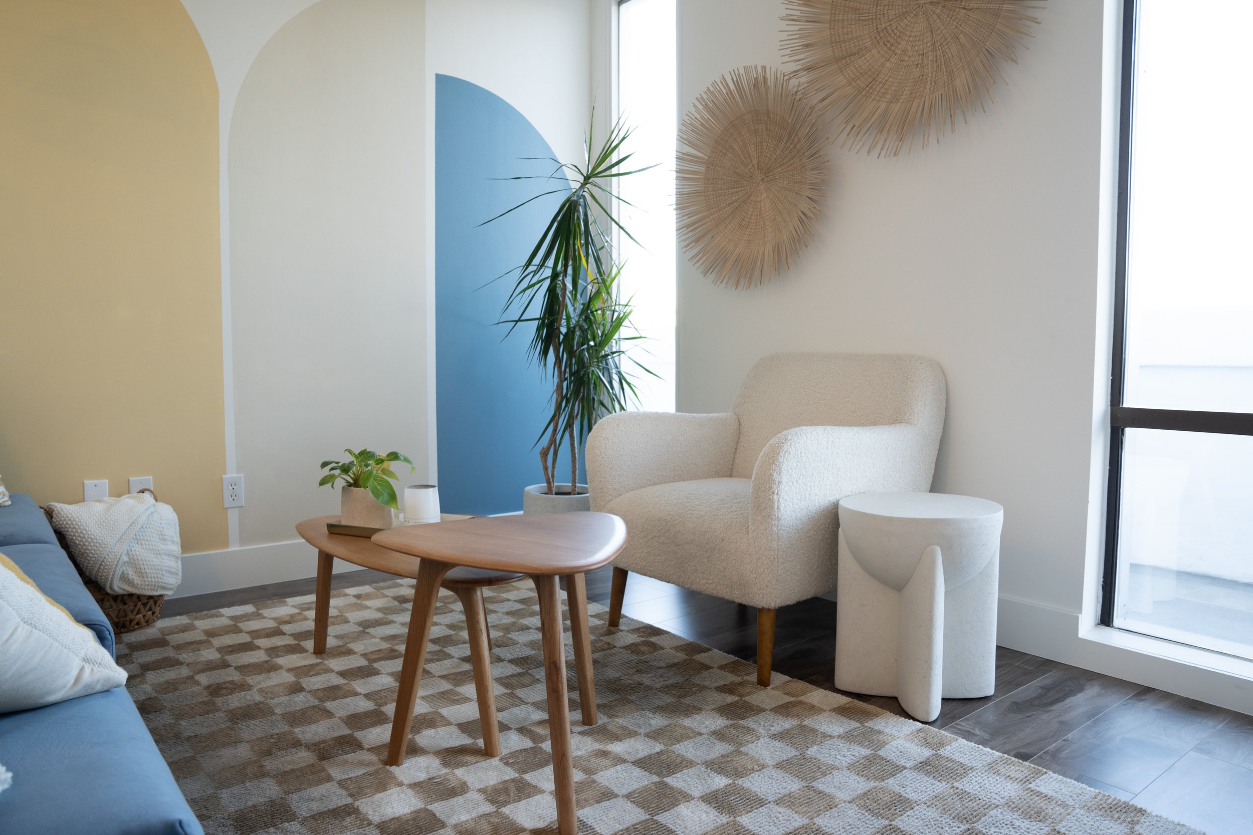 Living room with a white armchair, a small marble side table, a wooden coffee table with potted plants, and wall decor made of natural wooden slats. A large window lets in natural light, and a patterned rug covers the floor.