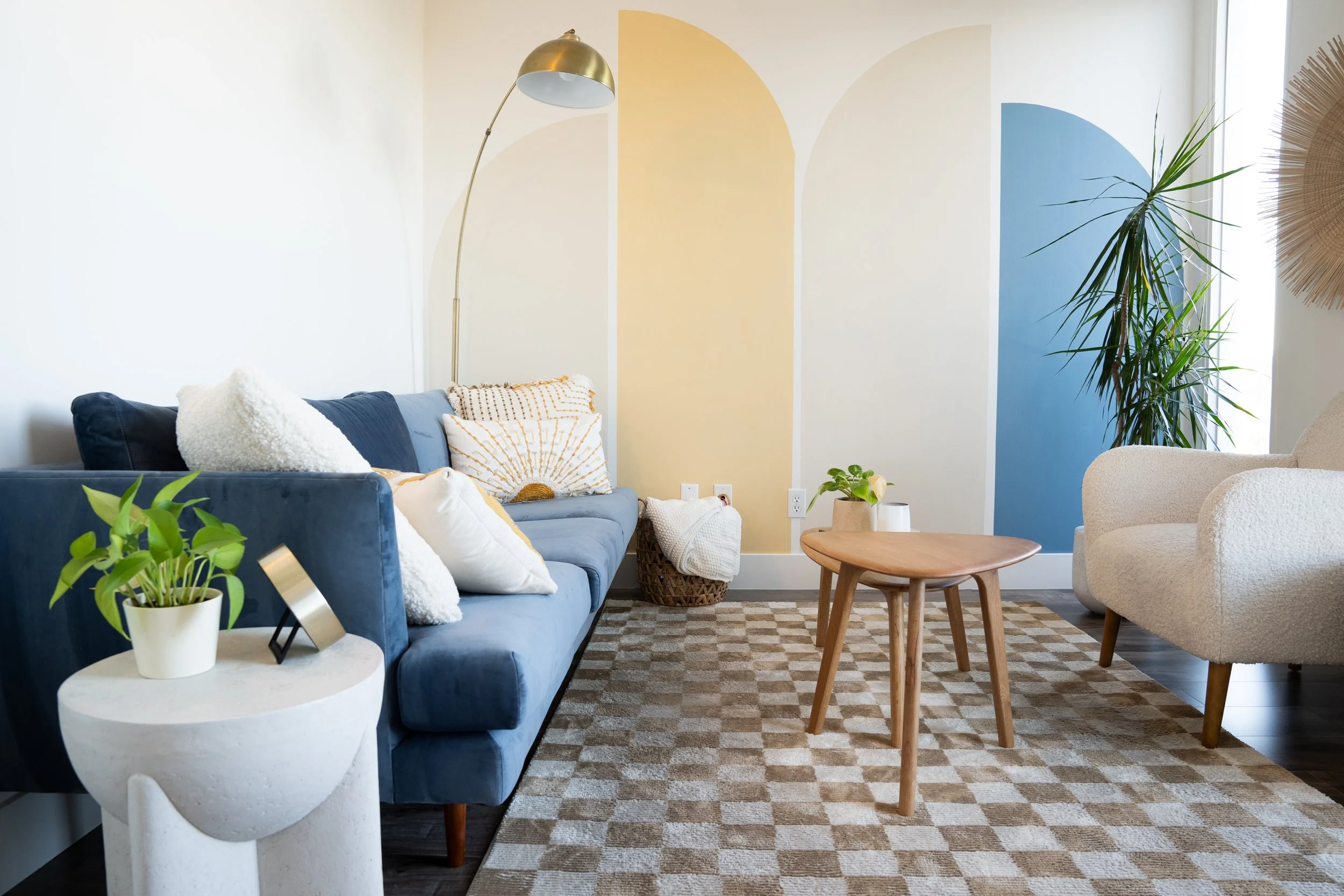 Living room with a blue sofa, white armchair, wooden coffee table, and various plants, with a colorful wall design in the background.