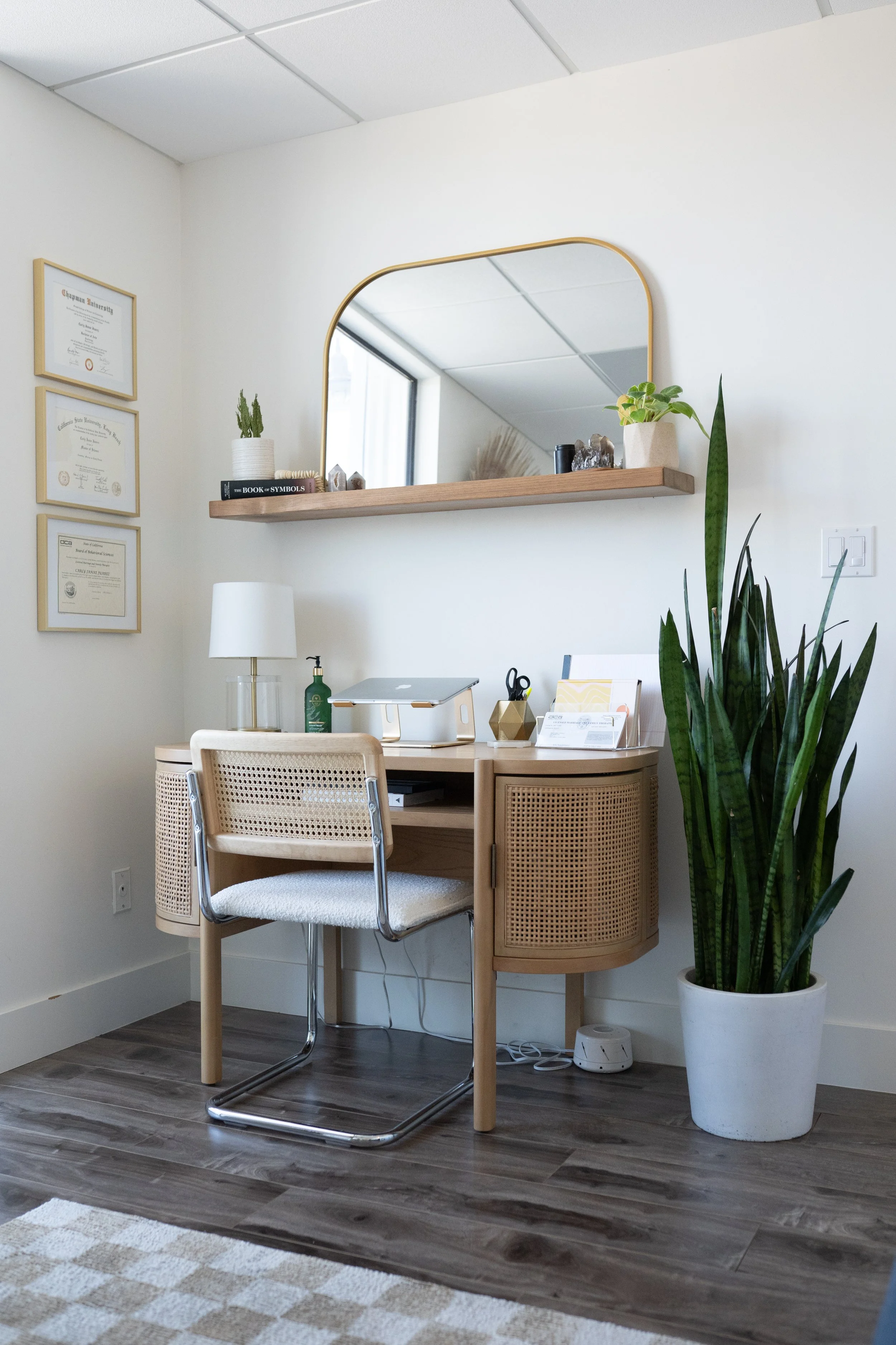 A home office setup with a small wooden desk, a white chair with a metal frame, a desk lamp, a potted snake plant, and wall decor including framed certificates and a wall mirror