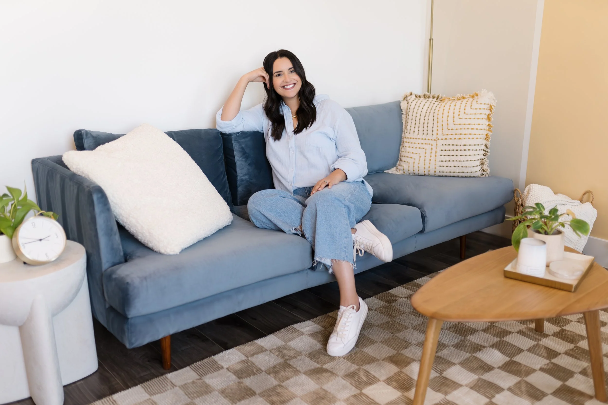 A woman with long dark hair sitting on a blue velvet sofa in a living room, smiling at the camera, wearing a light blue shirt and light blue jeans, with white sneakers. The sofa has three pillows, one white, one dark blue, and one with a geometric pattern. There is a wooden coffee table with potted plants and a tray nearby, and a small side table with a clock and a plant.