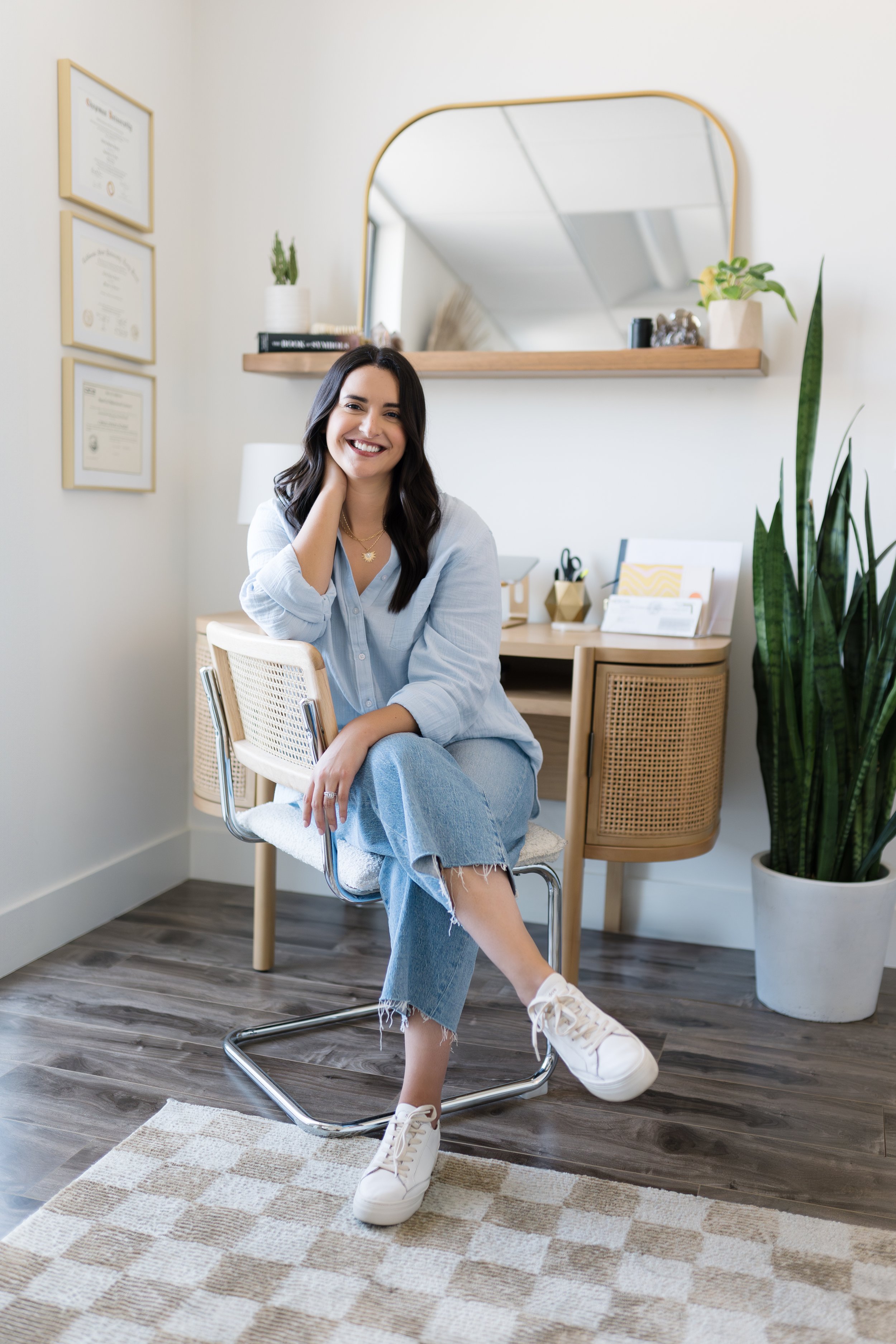 A woman with dark hair and a white shirt smiling and sitting on a chair in a modern office or home office decorated with plants, framed certificates, and a large mirror.