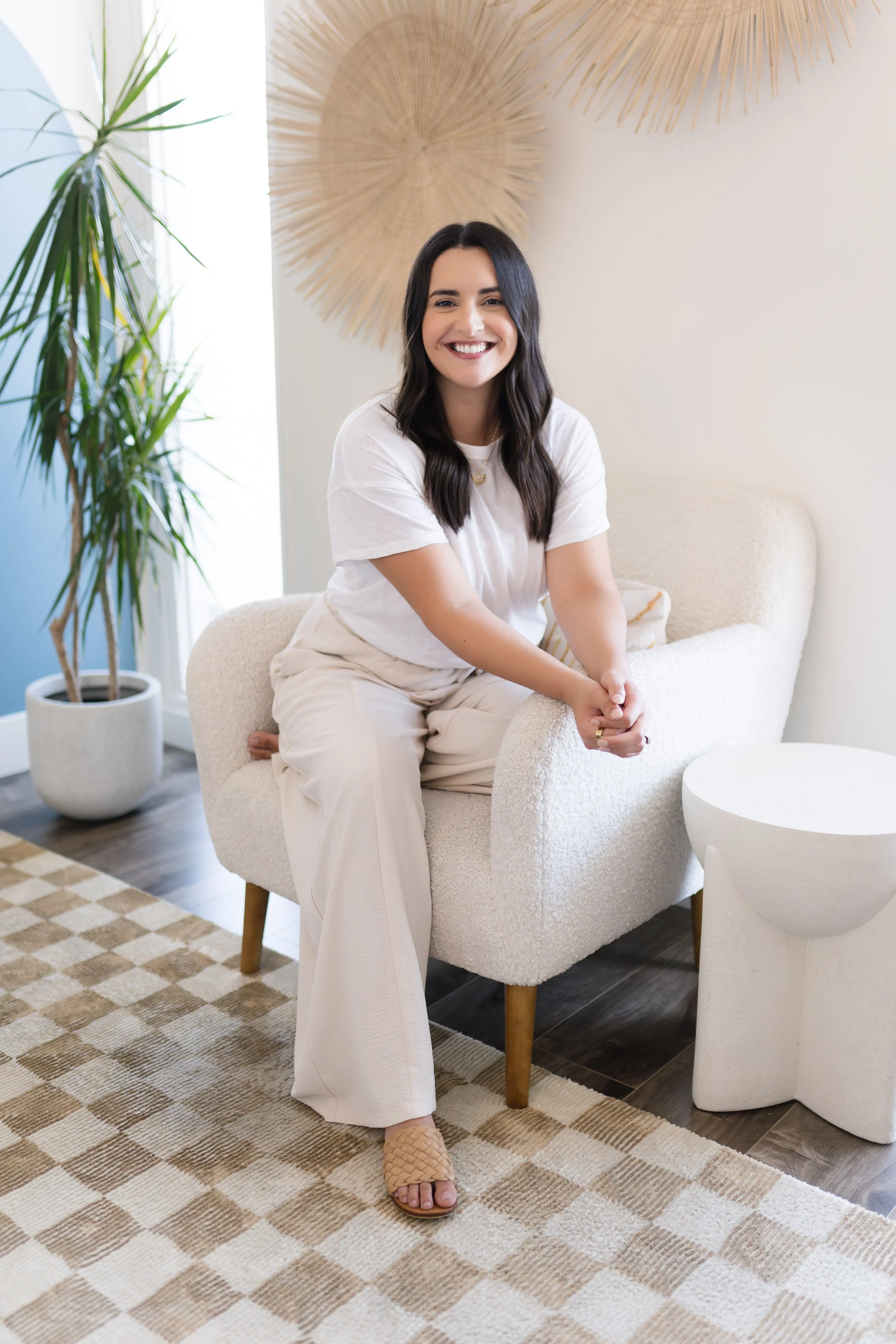 A woman with dark hair, smiling, sitting on a light-colored armchair in a modern interior, with a potted plant nearby and decorative wall art.
