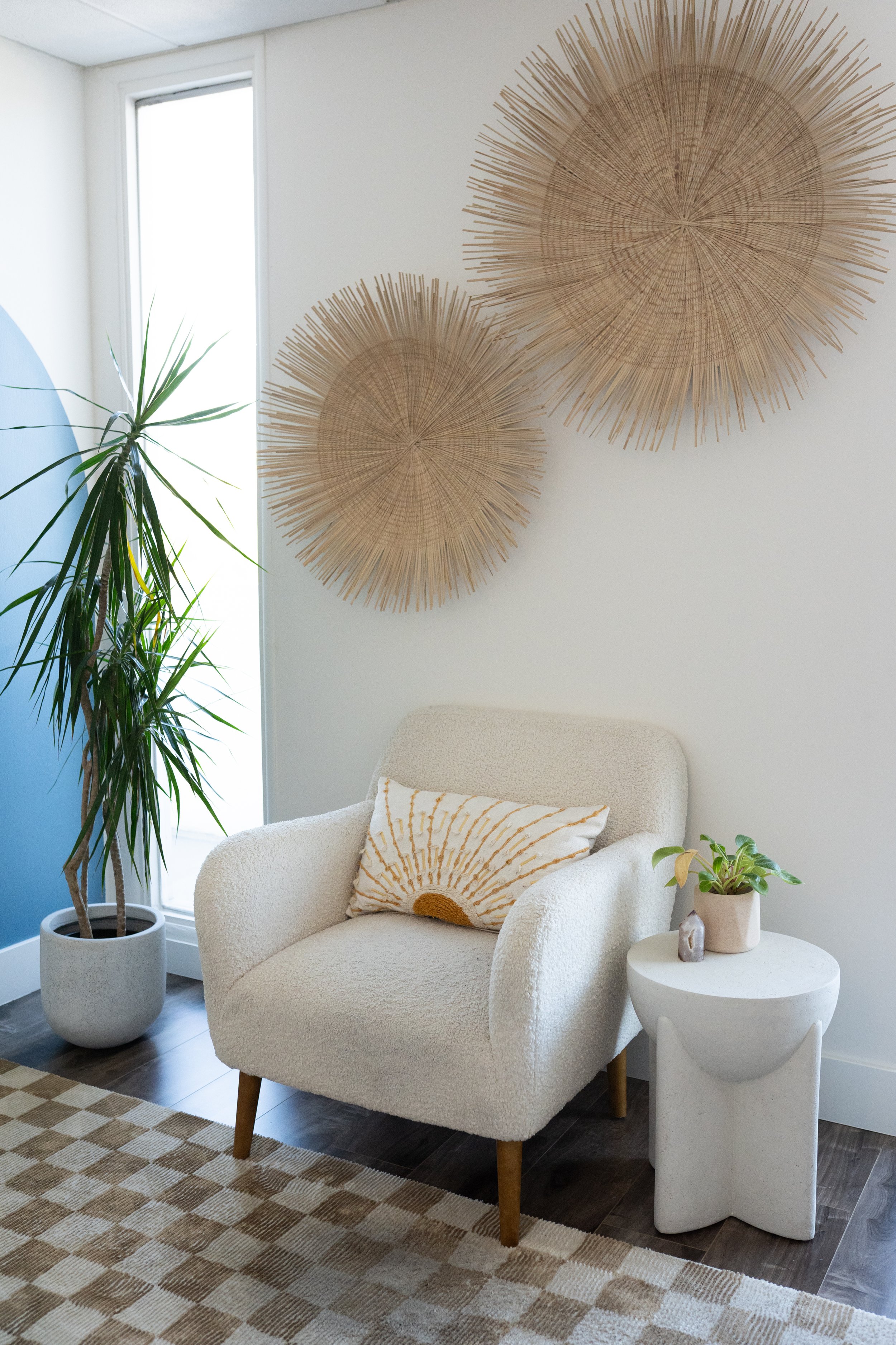 A cozy corner with a white upholstered armchair with wooden legs, a decorative pillow, a small round side table with a potted plant, large woven wall art, a tall green houseplant, and a window with sunlight.
