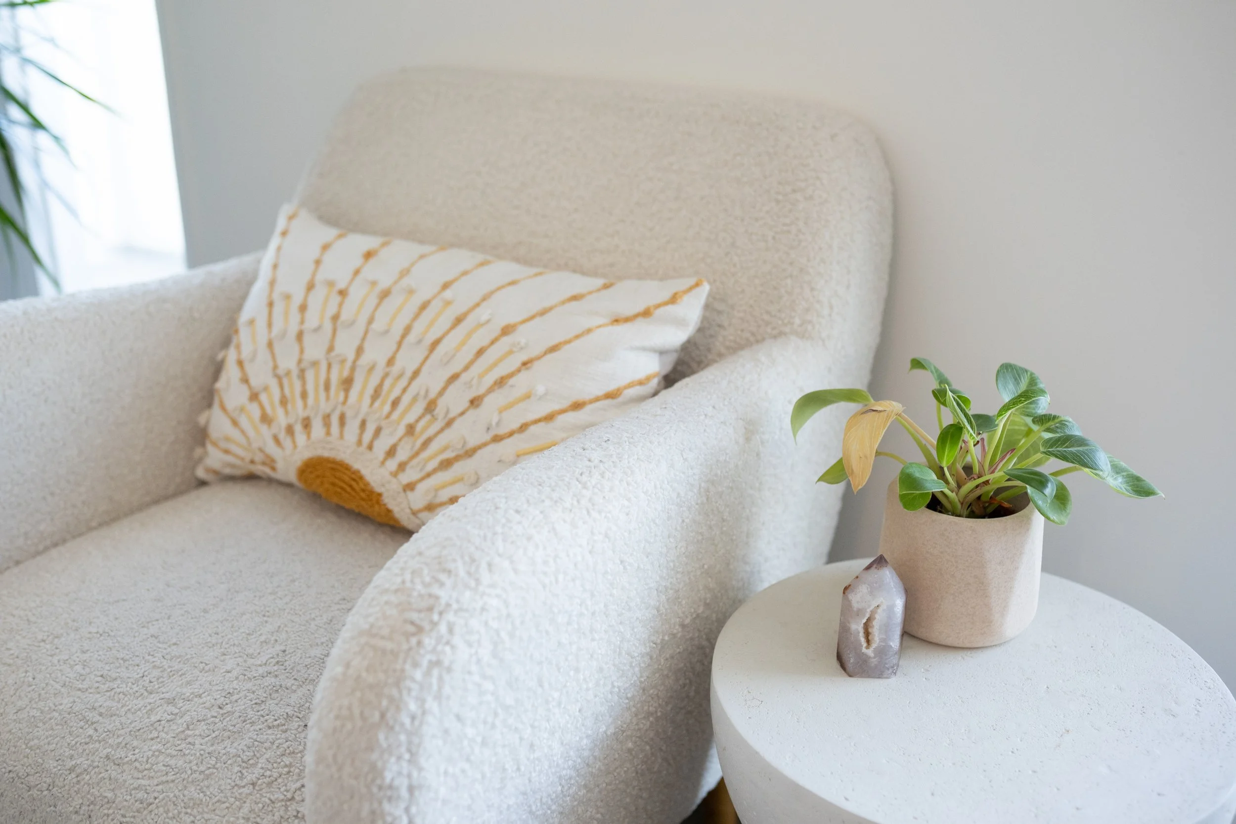 A cozy living room corner with a beige armchair, a decorative pillow with a sunburst design, a round side table with a potted plant, a small geode, and a window with natural light.