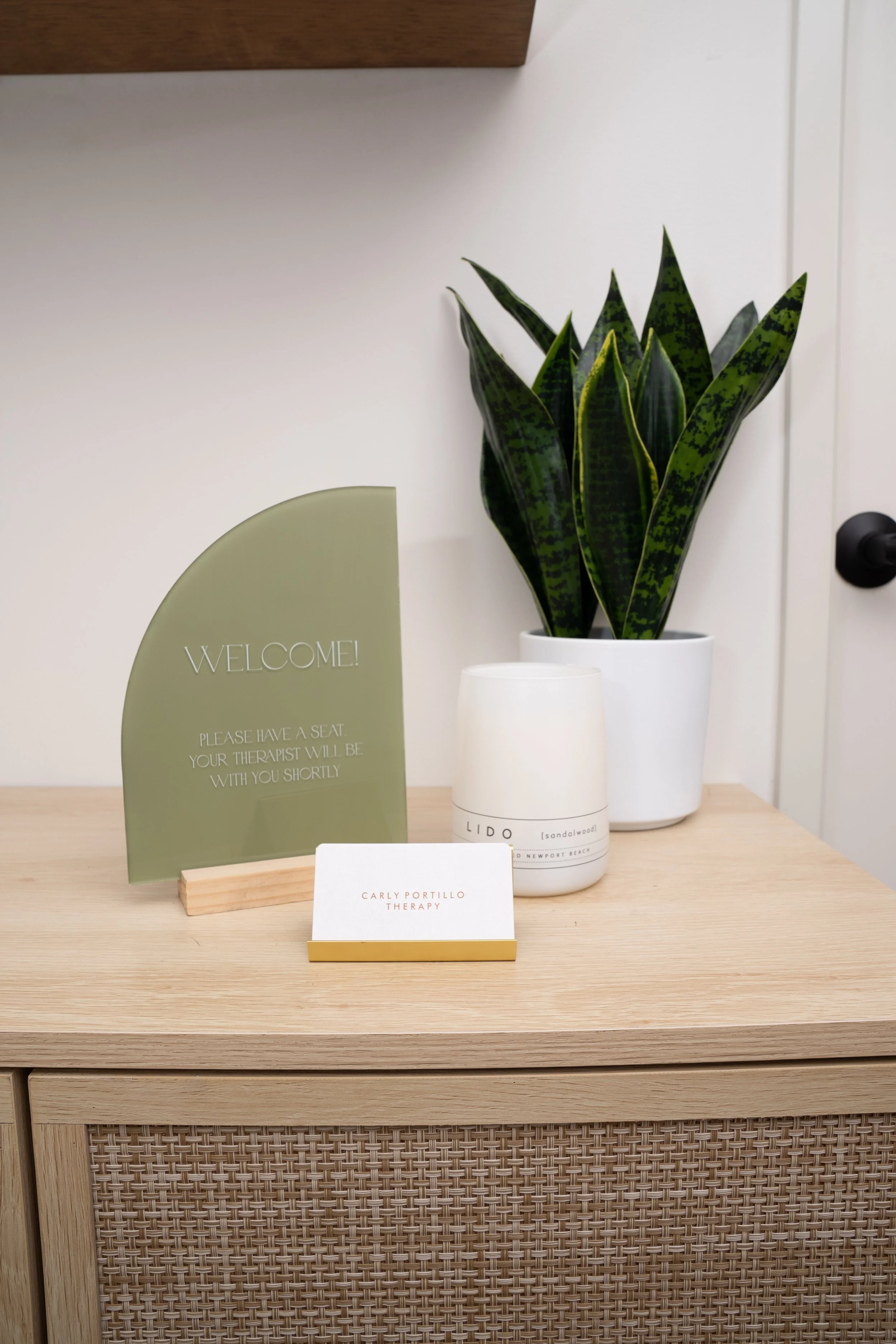 A wooden reception desk with a potted snake plant, a white candle labeled 'LIDO sandalwood,' a small white card with 'Carly Portillo Therapy' on it, and a green sign that says 'Welcome! Please have a seat. Your therapist will be with you shortly.'
