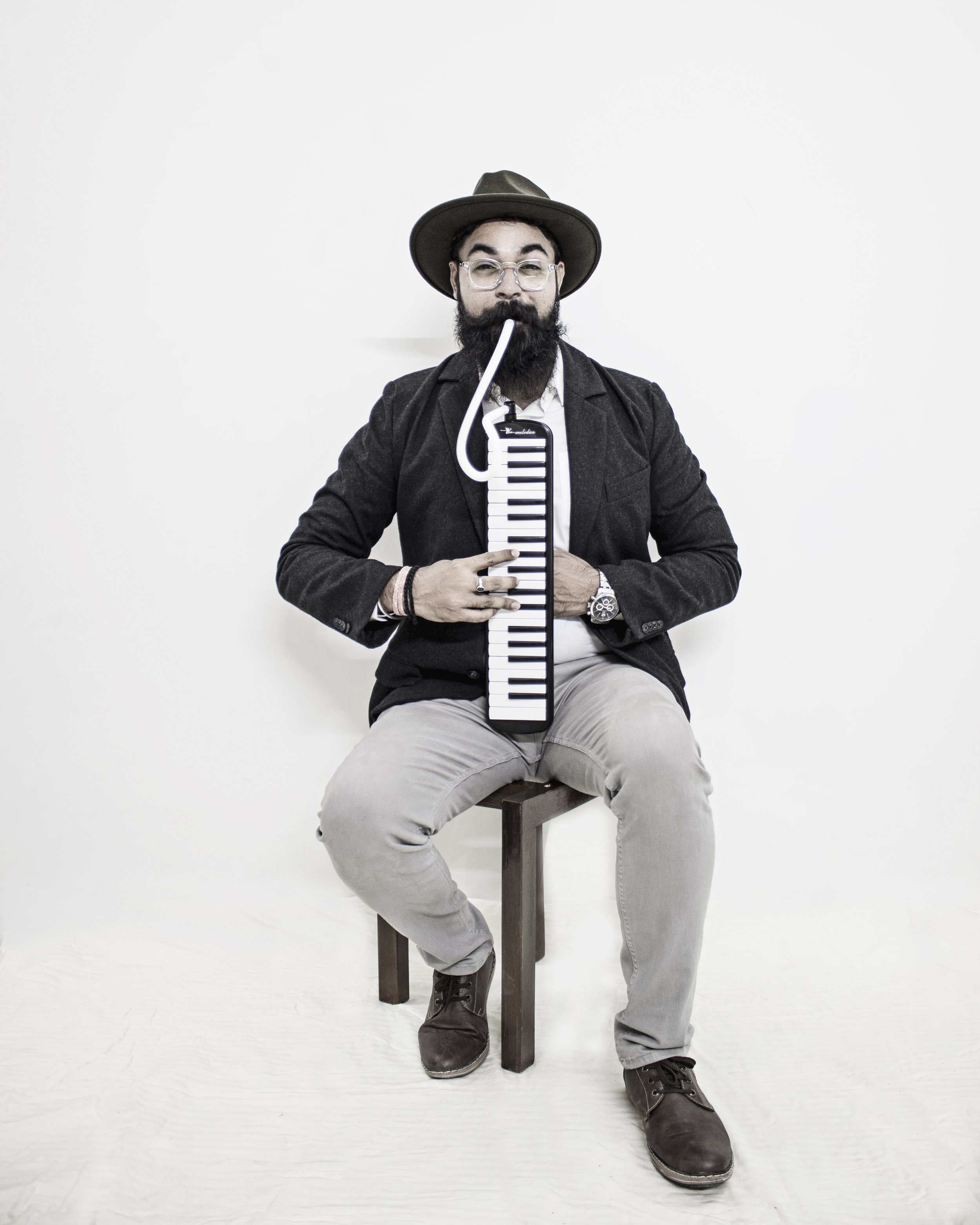 A man with glasses, a beard, and a hat sitting on a stool, holding a small keyboard and a musical note shaped like a tube in his mouth, against a plain white background.