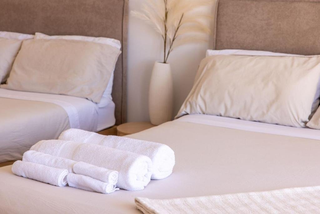 A neatly made bed with white linens and several rolled white towels placed on top, next to another bed with similar bedding, in a hotel room with a beige headboard and a white vase with tall dried plants on a small side table.