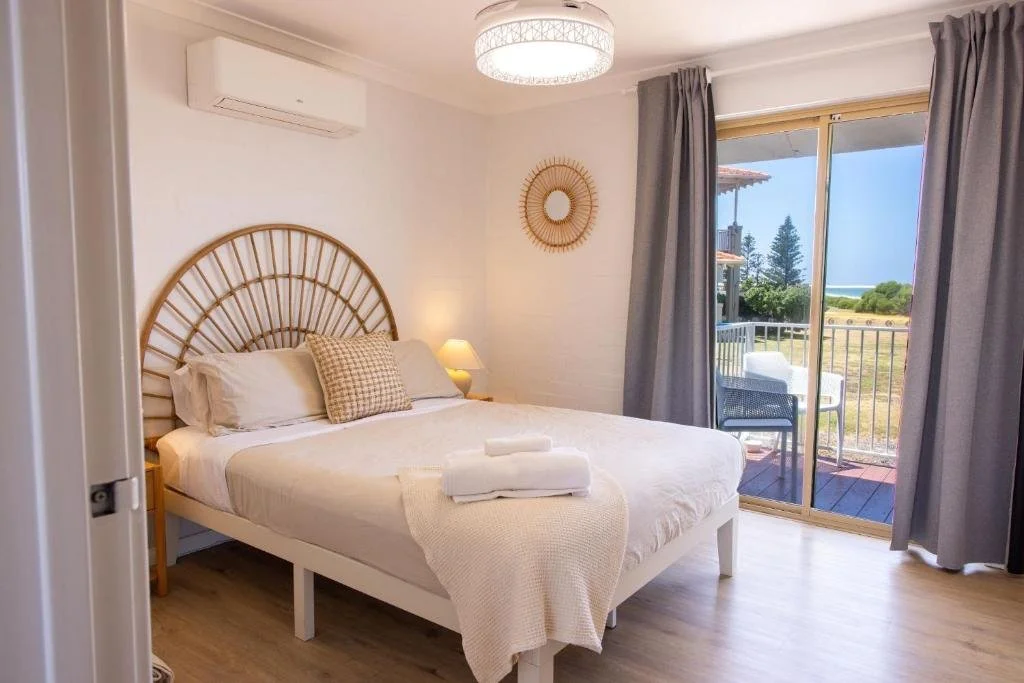A cozy bedroom with a large bed, beige bedding, and pillows, next to a sliding glass door leading to a balcony with outdoor chairs. The room has a white brick wall, curtains, a reading lamp, and a decorative mirror on the wall.